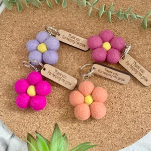 Könnte beinhalten: Vier blütenförmige Schlüsselanhänger in Lila-, Rosa- und Pfirsichtönen, jeder mit einem gelben Mittelpunkt. Die Schlüsselanhänger haben ein Holzschild mit der Aufschrift "Thank you for helping me grow."