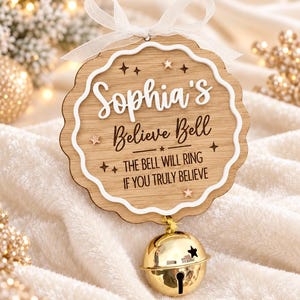 May include: A round wooden ornament with a scalloped edge, featuring the white text Sophia's Believe Bell and the black text THE BELL WILL RING IF YOU TRULY BELIEVE. It is decorated with small stars and a gold jingle bell hanging from a white ribbon.