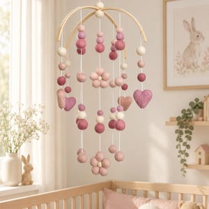 May include: A hanging mobile featuring a light wood arched frame with suspended strings of felt balls in shades of pink, cream, and mauve. The mobile includes felt flower and heart shapes interspersed among the round felt balls on white strings.