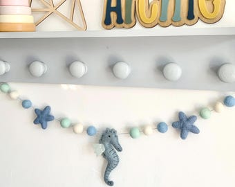May include: A decorative shelf with a wooden name sign that reads "Archie" in blue, green, and yellow. A garland of blue felt stars, seahorses, and light blue, green, and white felt balls hangs below. A wooden stacking toy and star are on the shelf.