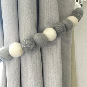 May include: A gray felt ball curtain tieback. The tieback is made of a series of gray and white felt balls. The tieback is holding back a light gray curtain. The curtain is hanging on a silver hook.