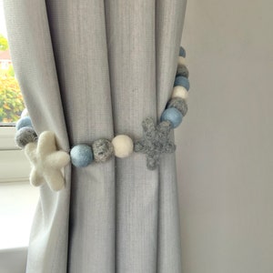 May include: A gray curtain with a white and blue felt ball garland with star shapes. The garland is used as a curtain tieback.