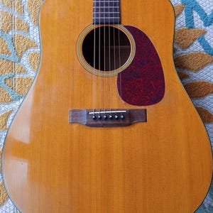 Puede incluir: Guitarra acústica con acabado de madera natural. La guitarra presenta una boca redonda, un golpeador rojo oscuro y seis cuerdas. El puente y el diapasón son de color marrón oscuro. El cuerpo es de color marrón claro.