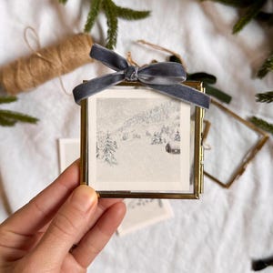 Könnte beinhalten: Ein kleiner, quadratischer, goldfarbener Metallrahmen mit einem Winterlandschaftsdruck. Der Druck zeigt eine verschneite Szene mit Bäumen und einer kleinen Hütte. Oben ist ein graues Samtband gebunden. Dekorativer Weihnachtsschmuck.