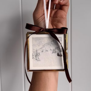 May include: A square glass ornament with a thin gold metal frame, featuring a black and white winter landscape illustration of snow-covered trees and a small cabin. The ornament is suspended by a white ribbon and adorned with a dark brown satin bow.