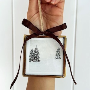 May include: A square glass ornament with a thin brass frame containing a white print of two black pine trees. The ornament is suspended by a natural jute twine loop and features a decorative dark brown satin ribbon tied at the top.