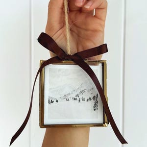 May include: A square glass frame with a thin gold metal border containing a black and white mountain landscape illustration. The frame is suspended by a natural jute twine loop and is decorated with a dark brown satin ribbon tied in a bow.