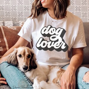 May include: White t-shirt with the words "dog lover" in a retro font. A long-haired dachshund dog is resting on a person's lap. The dog has tan and white fur. The person is wearing blue jeans.