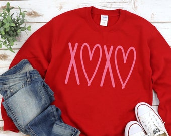 May include: Red sweatshirt with a pink "XOXO" graphic featuring two hearts. The sweatshirt is paired with blue jeans and white sneakers.
