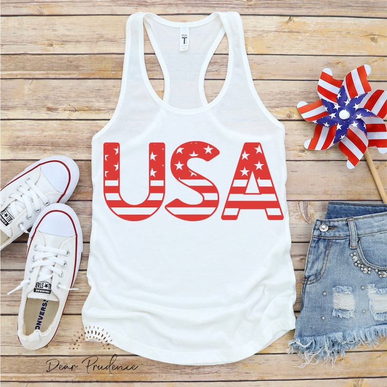 Puede incluir: Camiseta blanca de tirantes con la palabra "USA" impresa en rojo con un patr&oacute;n de bandera americana rojo, blanco y azul. La camiseta est&aacute; sobre una superficie de madera con un par de zapatillas blancas, unos pantalones cortos de mezclilla azul y una veleta roja, blanca y azul.