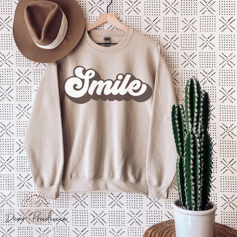 May include: A beige sweatshirt with a white retro-style "Smile" graphic. The sweatshirt is photographed against a white background with a black geometric pattern.