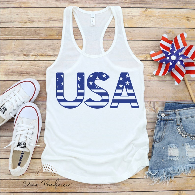 Puede incluir: Camiseta blanca de tirantes con la palabra "USA" impresa en azul y rojo, con estrellas y rayas. Las letras est&aacute;n estilizadas para parecerse a la bandera estadounidense.