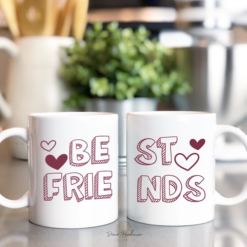May include: Two white ceramic mugs with a red outline of the words "BE" and "FRIENDS" on one mug and "BEST" on the other mug. Both mugs have small red hearts.