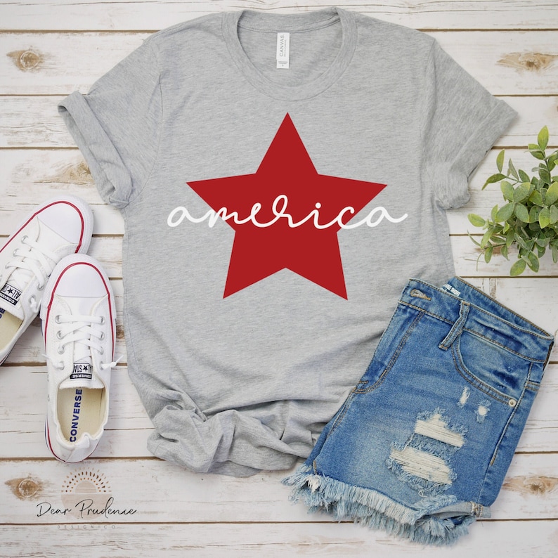 Op de afbeelding: Een grijs T-shirt met een rode ster en het woord "america" in wit schrift. Het shirt ligt op een houten oppervlak met een paar witte sneakers en een spijkerbroek.
