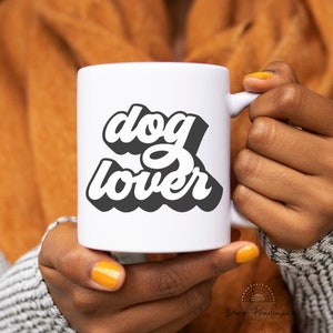 May include: A white ceramic mug with the words "dog lover" in a black and white retro font. The mug is held by a person with orange nail polish. The mug has a handle on the right side.