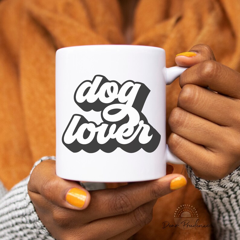May include: A white ceramic mug with the words "dog lover" in a black and white retro font. The mug is held by a person with orange nail polish. The mug has a handle on the right side.