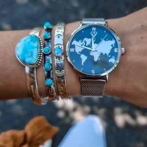May include: A silver watch with a black face and a world map design. The watch has a mesh metal band and is worn on a wrist with several silver bracelets with turquoise stones.