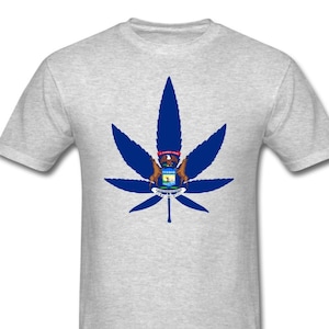 May include: A light grey t-shirt with a large blue cannabis leaf design. The leaf incorporates the Michigan state seal. The shirt is made of a soft material and has short sleeves.