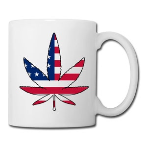 May include: White ceramic mug featuring a cannabis leaf design. The leaf is colored with the American flag, showcasing red and white stripes and a blue field with white stars. The mug has a curved handle.