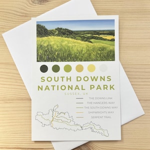 May include: A postcard depicting the South Downs National Park in Sussex, UK, with a landscape image and a trail map. The card includes a colour palette and the text "South Downs National Park". It rests on a wooden surface.
