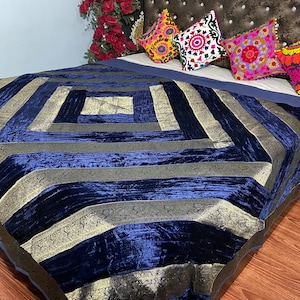 Op de afbeelding: Een donkerblauwe fluwelen deken met gouden geborduurde rand. De deken ligt gevouwen op een bed met decoratieve kussens.