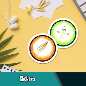 Könnte beinhalten: Zwei runde Aufkleber mit geometrischen Designs. Einer zeigt eine Flügelgrafik innerhalb eines orangefarbenen und weißen Randes. Der andere hat ein Pfeildesign innerhalb eines grünen und weißen Randes. Das Wort "Stickers" steht unten.