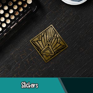 Op de afbeelding: Een vierkante gouden sticker met een geometrisch art deco-ontwerp. De sticker heeft een gestileerd gebouw met stralende lijnen en het woord "Stickers" onderaan.