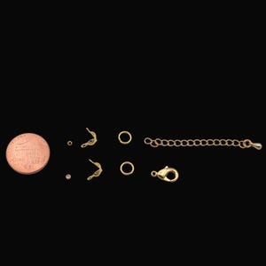 May include: A collection of gold-colored jewelry findings, including clasps, jump rings, and chain extenders, are arranged on a black background. A US one-cent coin is included for size reference. These components are used for jewelry making.