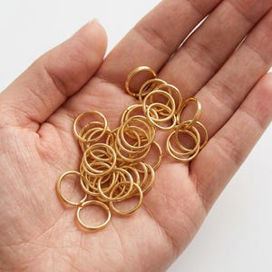 May include: A collection of small, circular gold-colored metal jump rings. These jewelry findings are thin, round, and feature a split opening, commonly used for connecting components in jewelry making and various craft projects.