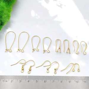 May include: A collection of gold-colored earring findings, including ear wires and fish hook styles, arranged on a white surface. The findings vary in size and design, with some featuring a loop for attaching beads or charms. A ruler is at the bottom.