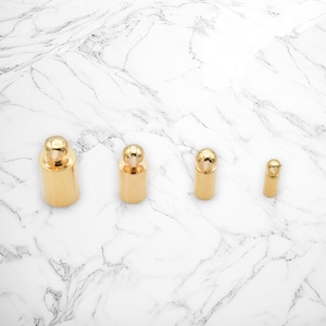 May include: Four gold-toned metal cord ends of varying sizes. The cord ends have a small loop at the top and a rounded ball at the bottom.