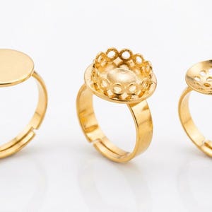 May include: Three gold-toned adjustable rings featuring different settings. The left ring has a flat circular plate, the center ring has a scalloped crown-style bezel setting, and the right ring has a concave circular plate with small perforations.