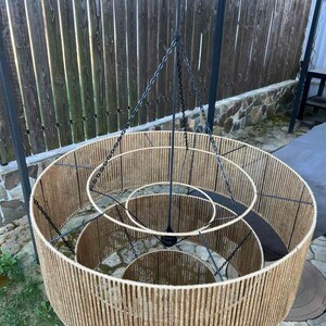 May include: A three-tiered chandelier featuring concentric circular frames wrapped in natural tan jute twine. The structure is supported by a central black metal rod and thin black chains that connect each tier to the main suspension point.