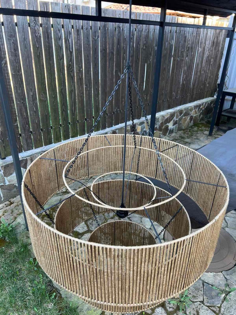 May include: A three-tiered chandelier featuring concentric circular frames wrapped in natural tan jute twine. The structure is supported by a central black metal rod and thin black chains that connect each tier to the main suspension point.