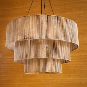 Peut inclure: Un luminaire suspendu à trois niveaux en corde naturelle tressée. Le luminaire est suspendu à un plafond avec un fond en bois.