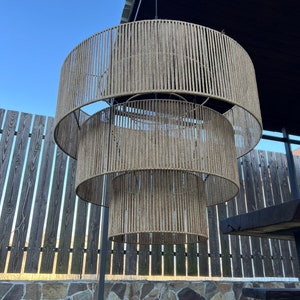 May include: A three-tiered pendant light fixture featuring cylindrical shades wrapped in natural tan jute rope. The structure consists of three concentric circular frames of varying diameters, suspended vertically by thin metal chains.