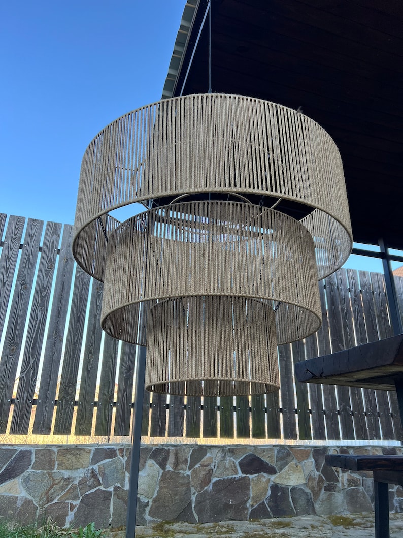 May include: A three-tiered pendant light fixture featuring cylindrical shades wrapped in natural tan jute rope. The structure consists of three concentric circular frames of varying diameters, suspended vertically by thin metal chains.