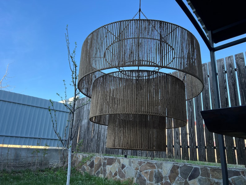 May include: A three-tiered drum chandelier featuring a brown rope-wrapped frame. The fixture consists of three concentric circular rings of varying diameters, each covered in vertical strands of brown cord, suspended by a central metal chain.