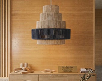 May include: A tiered pendant light fixture featuring a natural tan fiber wrap on the upper and lower sections, contrasted by a wide, dark navy blue band in the center. The cylindrical structure hangs from a thin black cord against a wood wall.