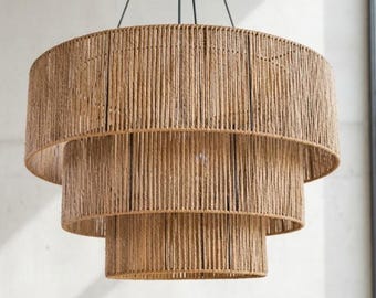 May include: A three-tiered pendant light fixture featuring cylindrical shades wrapped in natural tan jute rope. The structure is suspended by thin black wires connected to a central black circular ceiling mount, creating a layered, circular design.