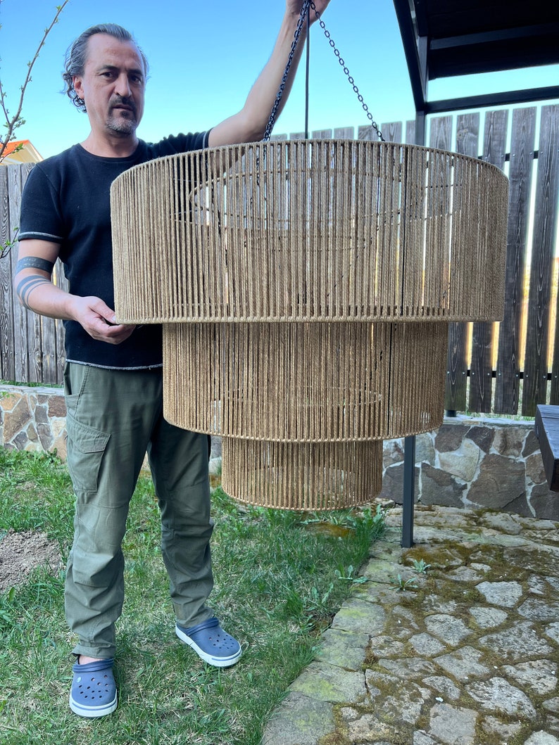 May include: A three-tiered cylindrical pendant light fixture constructed from a metal frame wrapped in natural tan jute twine. The fixture features three concentric circular tiers of varying diameters, suspended by a thin black metal chain.