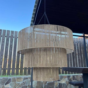 May include: A three-tiered cylindrical pendant light fixture featuring a frame wrapped in natural tan jute rope. The structure is suspended by a thin black cord and metal chain, creating a layered, circular silhouette with vertical rope detailing.