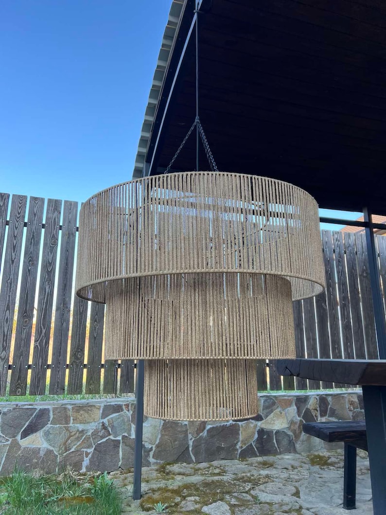 May include: A three-tiered cylindrical pendant light fixture featuring a frame wrapped in natural tan jute rope. The structure is suspended by a thin black cord and metal chain, creating a layered, circular silhouette with vertical rope detailing.