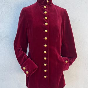 May include: A burgundy velvet jacket with a high collar and long sleeves. The jacket features a row of gold buttons down the front and on the cuffs. The jacket has a tailored fit and a classic design.