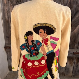 May include: Cream-colored jacket with embroidered figures of a dancing couple in traditional Mexican attire. The jacket features a black sombrero, red skirt, and colorful accents. The collar and cuffs have green stitching.