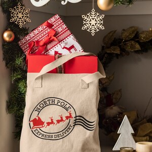 May include: A beige canvas tote bag with the words "North Pole Express Delivery" printed on it, filled with wrapped gifts. The gifts are wrapped in red and patterned paper. Christmas decorations, including ornaments and snowflakes, are in the background.