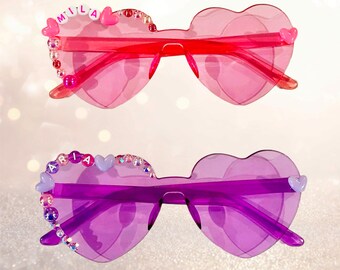 May include: Two pairs of heart-shaped sunglasses. One pair is pink with the name "MILA" spelled out in beads. The other pair is purple with the name "ARIA" spelled out in beads. Both pairs have decorative embellishments.