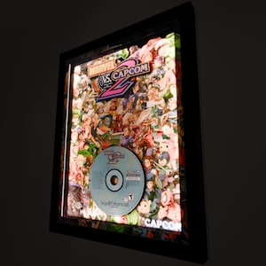 May include: A framed light-up display featuring the cover art for the video game "Marvel vs. Capcom 2" for the Sega Dreamcast. The display includes a clear plastic disc with the game's logo and a collage of characters from the game.