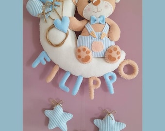 May include: A felt baby mobile featuring a teddy bear on a crescent moon, holding balloons. The mobile includes the name "FILIPPO" and hanging stars in blue and tan. A bow and the text "Io creazioni di Vale" are also visible.