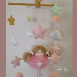 May include: A pink and white baby mobile with a felt doll, stars, and clouds. The doll is wearing a pink tutu and has brown hair. The mobile is hanging from a wooden frame.